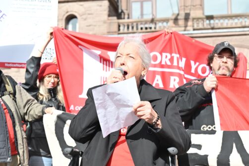 An older woman gives a speech at a rally with a North York Harvest Food Bank banner visible in the background.