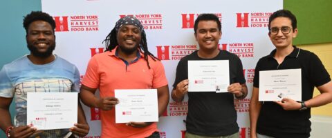 Four people stand in front of a North York Harvest Food Bank backdrop holding certificates for the Leadership in Logistics program.