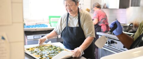 A woman in a kitchen wearing a hairnet moves a tray holding a large quiche, with people visible moving about in the background.