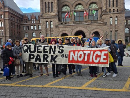 People stand outside Queen's Park in Toronto holding a large banner that says Queen's Park on NOTICE.