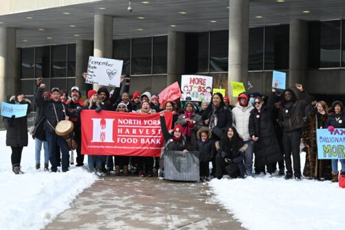 A large group of people stand outside at a rally at City Hall in Toronto. Many are holding protest signs about affordability, and there's a large banner for North York Harvest Food Bank.