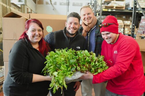 Four people smile as they all hold a large donation of fresh lettuce at North York Harvest Food Bank