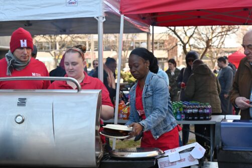 Three people wearing North York Harvest gear stand in front of BBQs preparing meat at an outdoor event.
