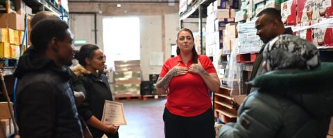 A woman wearing a red North York Harvest Food Bank t-shirt looks sincere, holding her hands to her chest, as she addresses students around her in a warehouse setting.