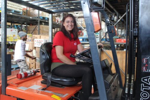 A woman in a red North York Harvest Food Bank shirt, smiles as she sits on a forklift inside a warehouse.