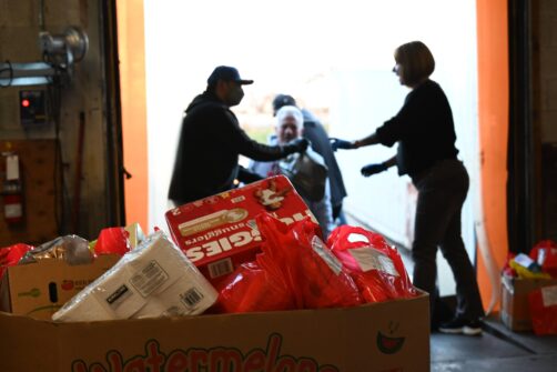 In the foreground is a box of donated items including food and diapers, in the background a person can be seen handing a bag of donated foods to another individual at the North York Harvest Food Bank warehouse.
