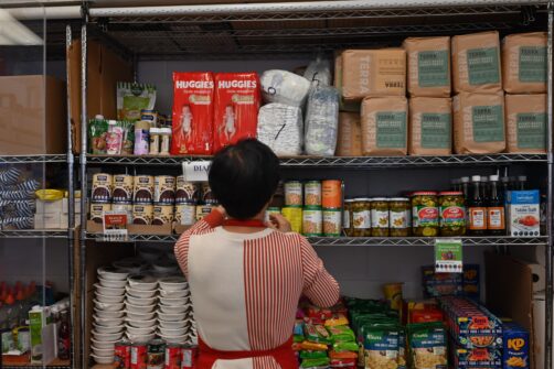 A woman is seen from behind, stocking full shelves of essential food and diapers at the North York Harvest Food Bank.