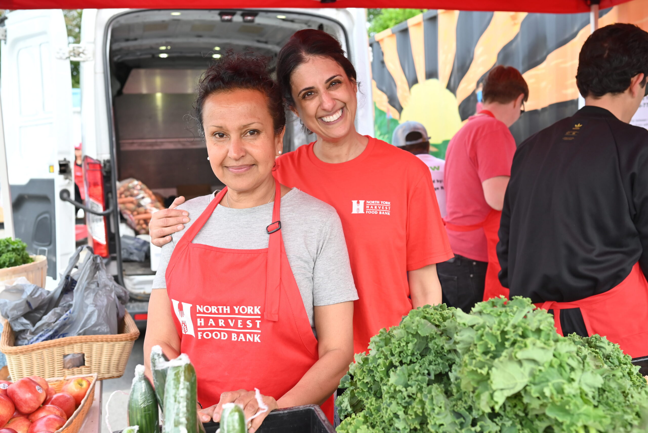 Two people wearing North York Harvest Food Bank aprons stand with their arms around one another smiling, in an outdoor market setting with fresh produce visible.