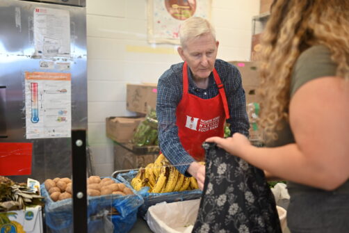 A man in a red North York Harvest Food Bank apron hands food to a woman holding a grocery bag at a food bank.