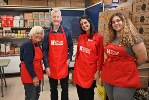 Four people wearing red North York Harvest Food Bank aprons smile in a food bank.
