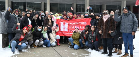 A group of people hold the North York Harvest Food Bank banner outside of City Hall, smiling, some with their fists in the air triumphantly.
