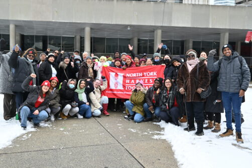 A group of people hold the North York Harvest Food Bank banner outside of City Hall, smiling, some with their fists in the air triumphantly.