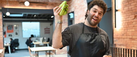 A chef stands in front of two boxes of produce, holding a bunch of bananas in the air, smiling.