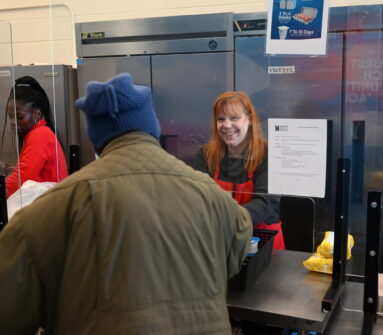 A woman in a North York Harvest Food Bank apron smiles while serving food to a community member at the food bank,