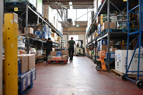 North York Harvest Food Bank's warehouse is seen buzzing with activity, people walk down the aisle as a person on a forklift drives by. They are surrounded by ceiling-high racking filled with non-perishable food items for the community.