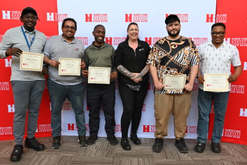 A group of people holding certificates stand in front of a North York Harvest Food Bank backdrop, smiling.
