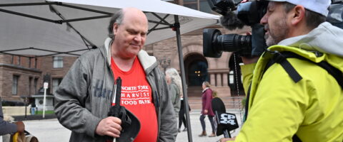 A person wearing a red North York Harvest Food Bank shirt who is blind is interviewed by a reporter holding a video camera outside Queen's Park in Toronto.