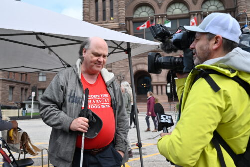A person wearing a red North York Harvest Food Bank shirt who is blind is interviewed by a reporter holding a video camera outside Queen's Park in Toronto.