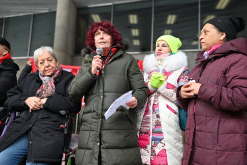 Four people in winter gear stand outside of City Hall during a rally in Toronto. One person with red hair is addressing the audience, holding a microphone.