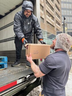 A person in a North York Harvest Food Bank jacket leans out of a delivery truck to pass a box of food to someone on the ground.