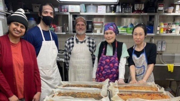 People in a kitchen setting stand in front of trays of prepared meals.