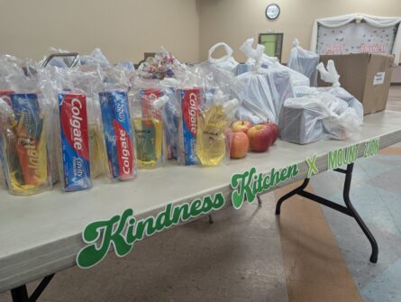 Care packages containing food and household items are arranged on a table with the words Kindness Kitchen & Mount Zion on it.
