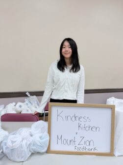 A young person stands in front of a table with care packages and a sign that reads Kindness Kitchen.