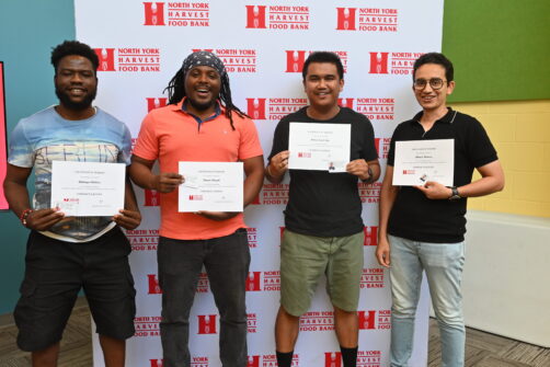 Four people hold graduation certificates, smiling in front of a North York Harvest Food Bank backdrop.