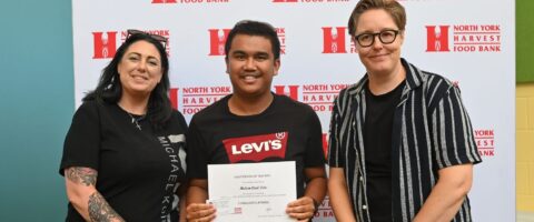 Three people smile for a photo in front of a North York Harvest Food Bank backdrop. The person in the centre is holding a Leadership in Logistics graduation certificate.