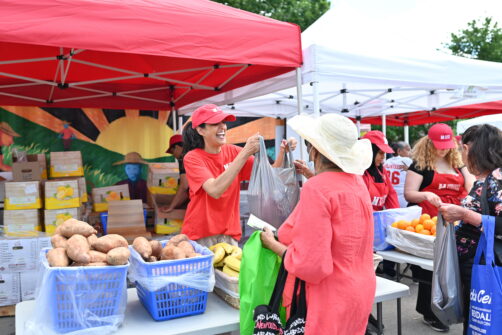 A person in a red North York Harvest Food Bank hat and shirt hands a bag of fresh vegetables to a community member at an outdoor produce market.