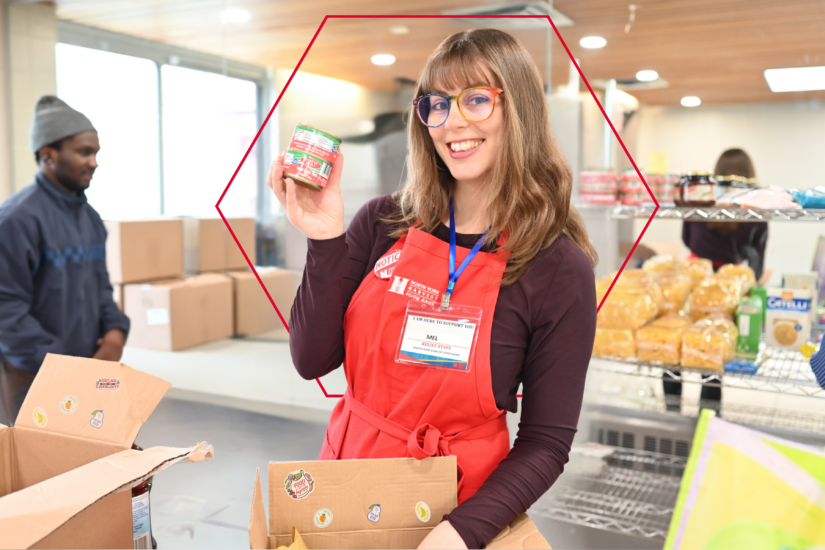 A woman in a red North York Harvest Food Bank apron holds up a can of food with boxes and shelves around her. In the background, other volunteers are working at the food bank.
