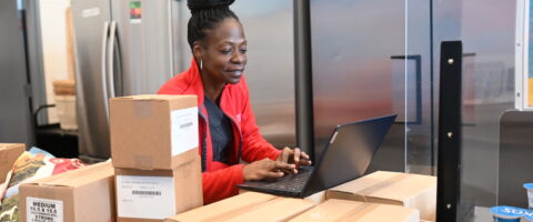 A person wearing a red North York Harvest Food Bank sweater types onto a laptop piled on boxes of food in a food space setting.