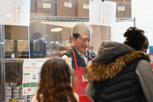 A person wearing a red North York Harvest Food Bank apron gives food to two clients, a mother and daugher. They are visible from behind to protect client privacy.